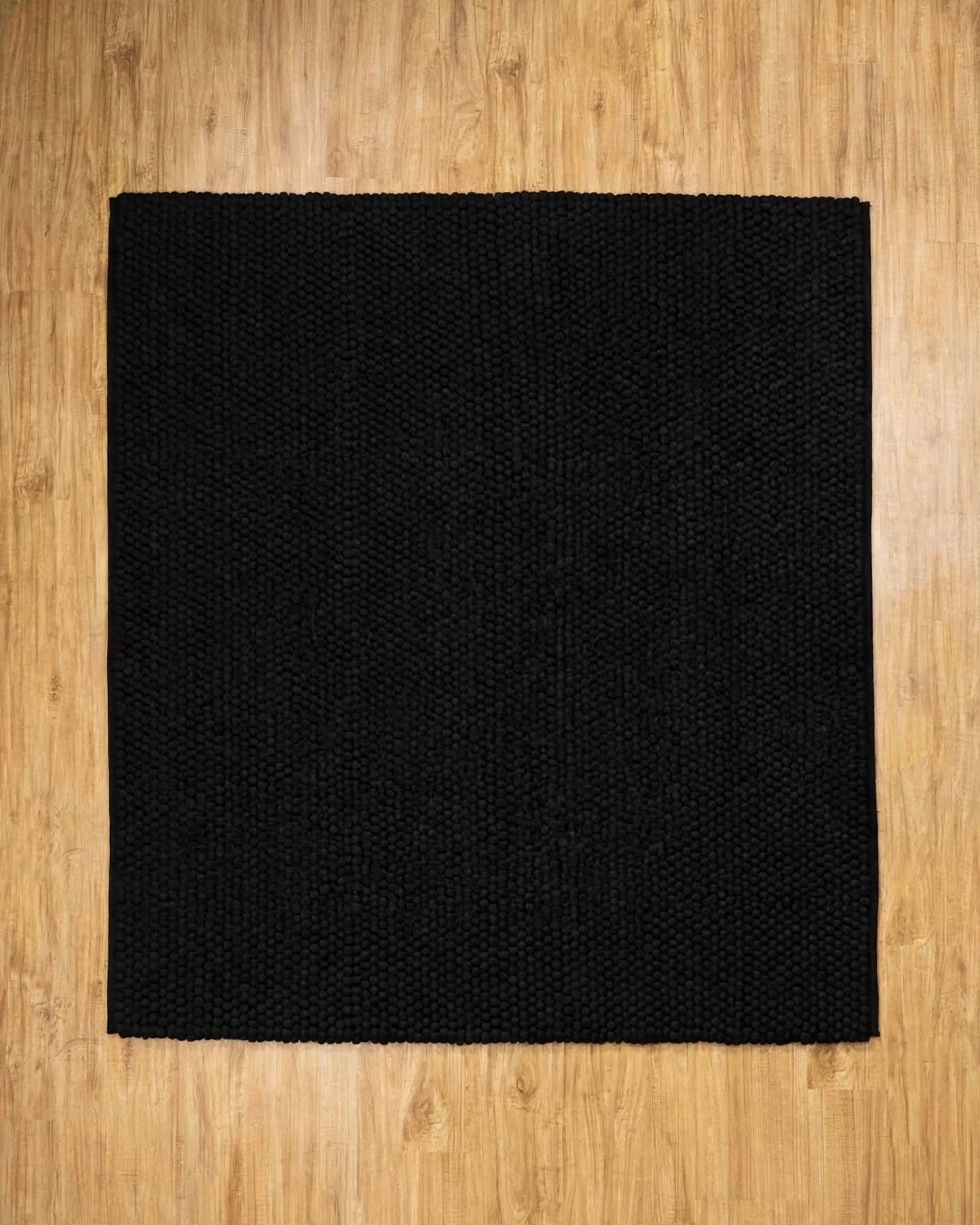 Charcoal Black Chunky Wool Area Rug, handmade in Bhadohi India – a square black woven rug laid flat on a light brown wood floor