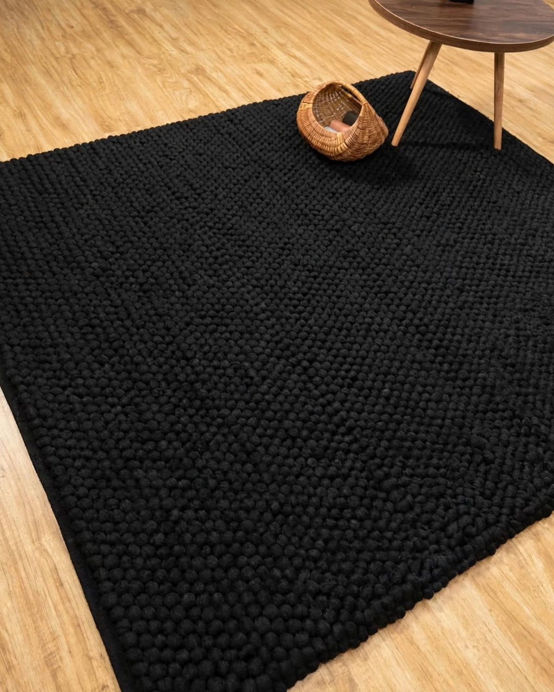 Charcoal Black Chunky Wool Area Rug, handmade in Bhadohi India – a large black textured area rug laid on a light wood floor, with a small woven